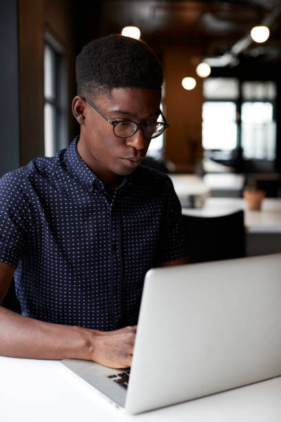 istock Young black male creative sitting at a desk in an open plan office using laptop, close up, vertical 1146471370