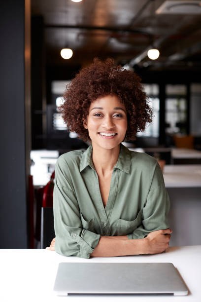 istock Millennial black female creative sitting at desk in an open plan office smiling to camera, vertical 1146471456