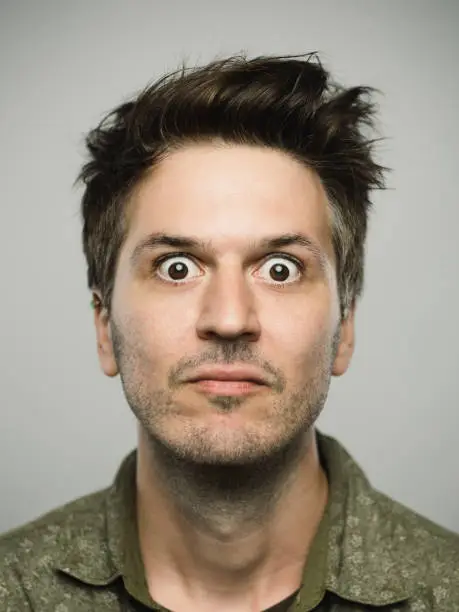 Close up portrait of young adult man with sursprised expression looking at camera against gray white background. Vertical shot of caucasian real people shocked in studio with brown hair and modern spiky haircut. Photography from a DSLR camera. Sharp focus on eyes.