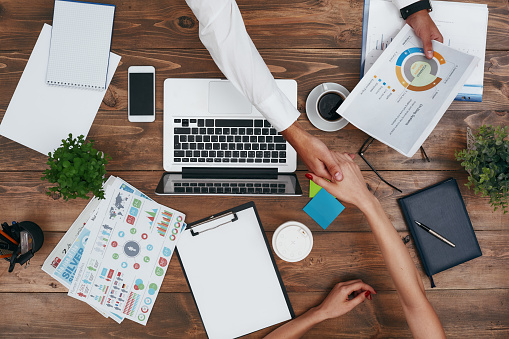 Top view of man and woman shaking hands while discussing new business strategy. Laptop, potted plant, diary, coffee cup, glasses, charts and other supplies on brown wooden desk. Colleagues working in the office. Horizontal shot