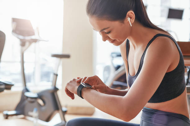 ergebnisse prüfen. seitenblick auf die schöne junge frau, die ihr sportarmband anschaut, während sie sich im fitnessstudio ausruhen - checking the time watch women looking stock-fotos und bilder