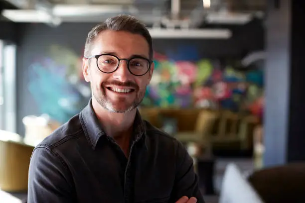 Photo of Middle aged white male creative in casual office lounge area looks to camera smiling, close up