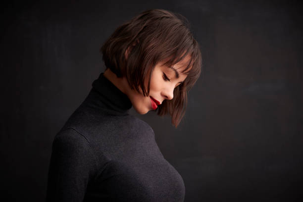 Close-up portrait shy smiling young woman wearing roll neck sweater while standing at dark background.
