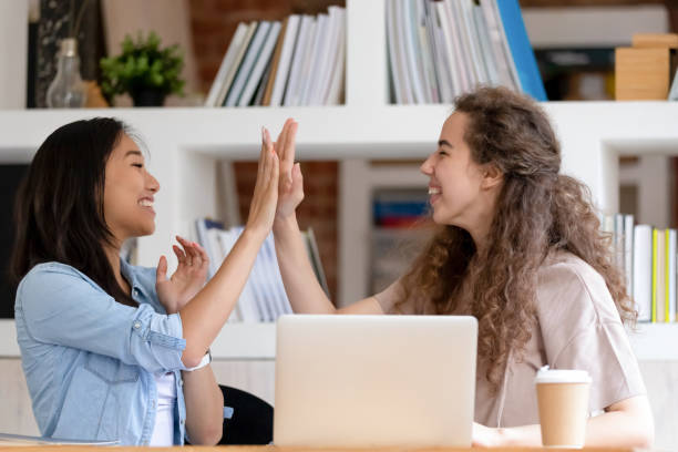 Cheerful student girls feels happy celebrating success giving high five Profile view cheerful asian and caucasian student girls sitting at table university library feels happy giving high five celebrate successful exam pass, getting scholarship, fruitful teamwork concept two groups stock pictures, royalty-free photos & images