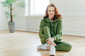 Horizontal shot of sporty red haired woman dedicated to fitness, wears green tracksuit and white sportshoes, poses on floor in spacious room, has rest after pilates exercises, yoga practice.