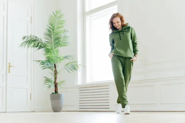 Photo of Full length shot of active pleased young woman with slim body, keeps hands in pockets of tracksuit, focused down, ready for doing cardio exercises, has determined look, pilates exercising indoor