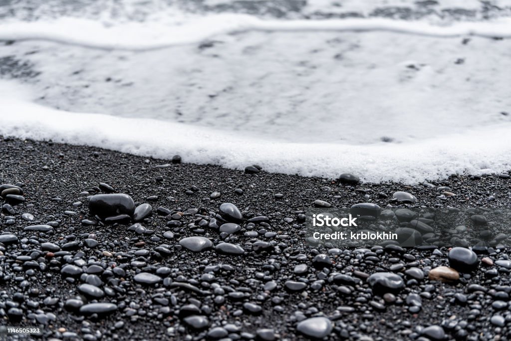 Black volcanic stones on sand beach in Reynisfjara, Iceland with water waves crashing on shore to shiny wet rocks in Vik Abstract Stock Photo