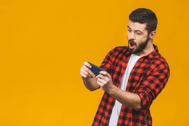 aufgeregter bärtiger mann im karierten hemd, der auf dem smartphone isoliert über gelbem hintergrund spielt. - portable player stock-fotos und bilder