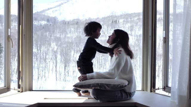 A child and his mother kissing near a window and watching snow falling in forest..