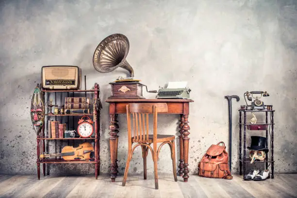 Photo of Antique chair, old typewriter, retro radio,  gramophone on wooden desk, books, clock, camera, binoculars, fiddle, keys on shelf, mask, cylinder hat, shoes, cane, backpack. Vintage style filtered photo