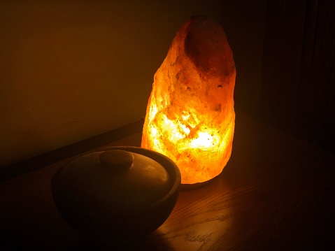 a himalayan rock salt lamp sits on a shelf next to a wooden bowl this photo is dark and moody with heavy shadow