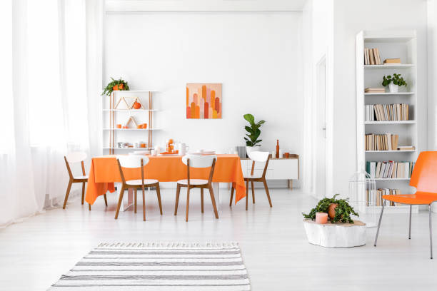 istock White flat interior with window with curtains, bookcase, rack with decor and long dining table with orange cloth and four chairs in the real photo 1146547237