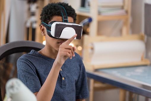 A teen boy sits in a chair wearing virtual reality glasses and gesturing as he plays a game.