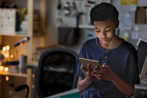 A teen boy plays a game in his room on a digital tablet at night.