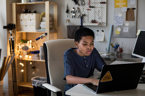 A serious teenage boy sits at a desk in his bedroom and types on a laptop computer.