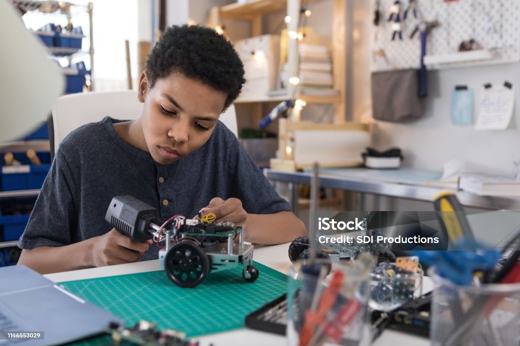 Teen boy solders wires to build robot A serious teen boy uses a soldering gun to connect wires as he builds a robot at home. Child Stock Photo