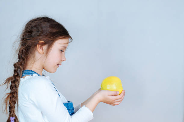 portrait émotionnel petite belle fille avec des tresses en jeans salopette mangeant des piqûres tenant une pomme. 6-7 ans studio - 6 7 years little girls child standing photos et images de collection