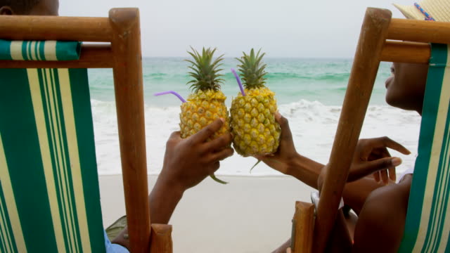 Rear view of African american couple toasting pineapple juice on the beach 4k