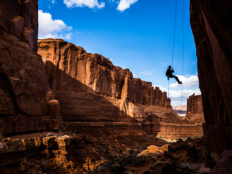A man hanging in mid air by a single rope is shadowed by the narrow canyon he is in, but behind him the red sandstone cliffs are washed in late evening light.