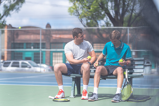 Two young male tennis players sit on the sideline bench and talk after a rousing match. They're sitting on an outdoor bench and both are smiling as one hands the other a ball.