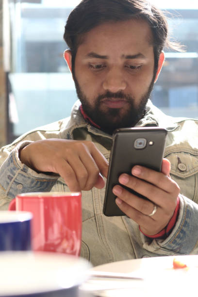 image of hindu indian man smiling, looking and reading text message on mobile phone at breakfast dining table, scrolling checking social media and surfing internet news on smart phone first thing in morning, mug of black coffee / cappuccino, hot chocolate - young adult reading newspaper the media imagens e fotografias de stock
