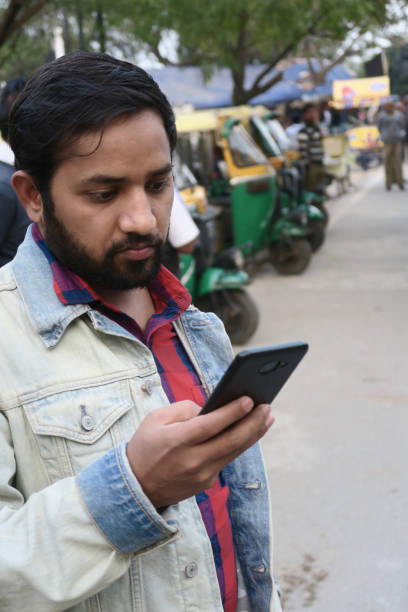Image of Hindu Indian man looking at phone screen reading text message / messaging and texting, holding mobile phone and checking social media / surfing Internet new information on smart phone device technology, auto rickshaws tuk tuks transport in India Stock photo of young man with beard looking at mobile phone screen, reading text message, scrolling fingers and messaging / texting, holding mobile phone, checking social media Internet. auto rickshaw taxi india stock pictures, royalty-free photos & images