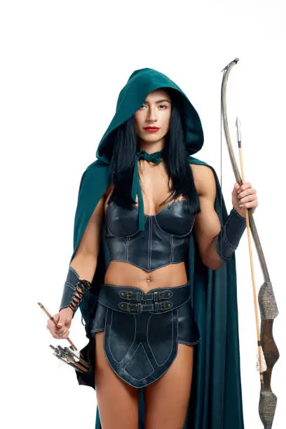 Beautiful and young girl making cosplay of fantasy creature. Gorgeous archery in emerald mantle with hood and leather armor posing at camera. Elegant woman holding bow and arrow and ready for fight.