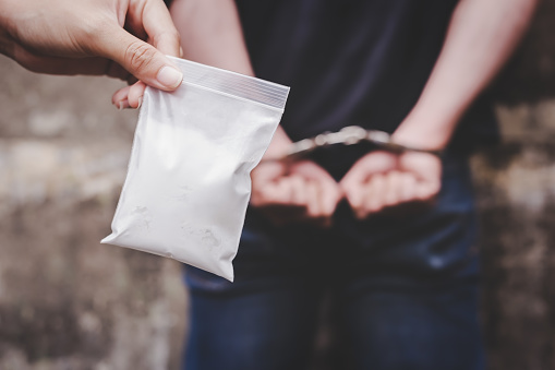 Drug dealers was under arrested and locked the arms bad guy behind his back at the wall by using handcuffs. It has evidence, policeman shows cocaine, heroine or narcotic contains in the plastic bag