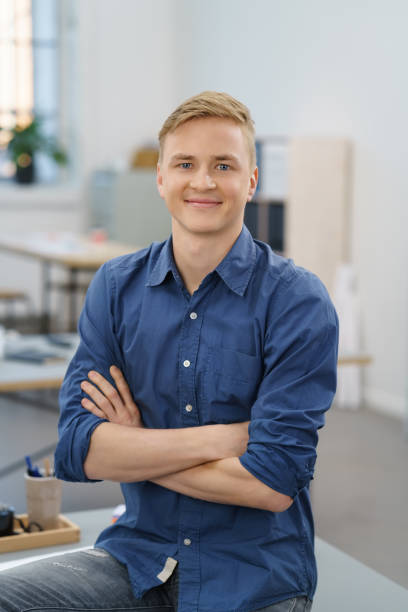 Confident young businessman with a friendly smile Confident young businessman with a friendly smile sitting on a table at the office with folded arms grinning at the camera blonde hair stock pictures, royalty-free photos & images