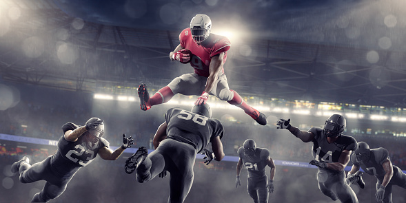 An American football player in mid air, jumping over the head of a rival player whilst holding onto the ball.The player is wearing a generic red and white football uniform and wear a helmet with a visor. Opposition players are dressed in black uniforms and are diving from all directions.
