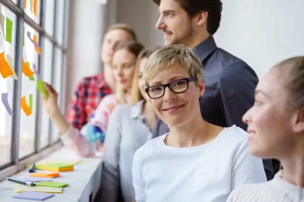 A portrait of a smiling woman wearing glasses in a happy team office environment while discussing ideas with sticky notes.