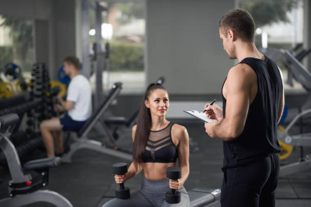 Energetic girl and man in black talking in gym. Heathy fit energetic girl training with coach in black sportswear in gym with some sports equipment. Young athlete helping woman standing and writing in tablet. personal trainer cost stock pictures, royalty-free photos & images