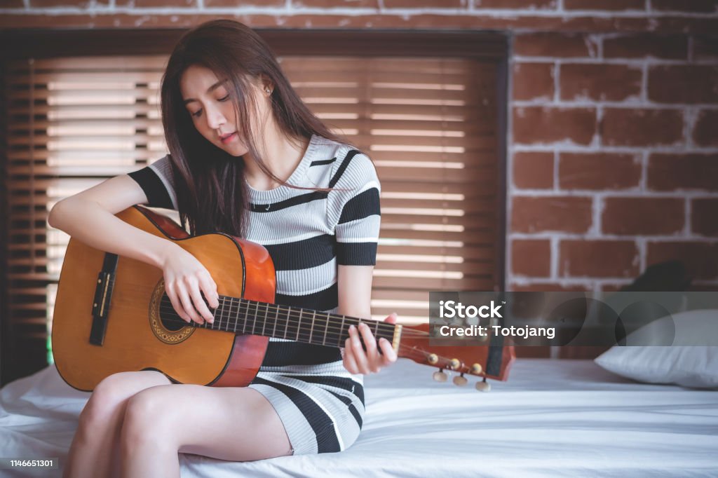 Young beautiful Asian woman is playing acoustic guitar on the bed at her bedroom, vintage warm tone Acoustic Guitar Stock Photo
