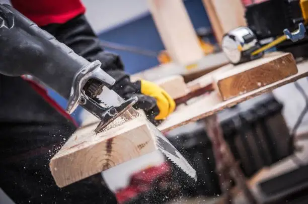 Small Construction Project. Men Cutting Piece of Wood Board Using Reciprocating Saw. Construction Site Power Equipment.