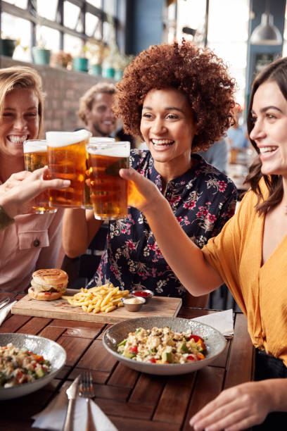 Four Young Female Friends Meeting For Drinks And Food Making A Toast In Restaurant Four Young Female Friends Meeting For Drinks And Food Making A Toast In Restaurant drinking beer stock pictures, royalty-free photos & images