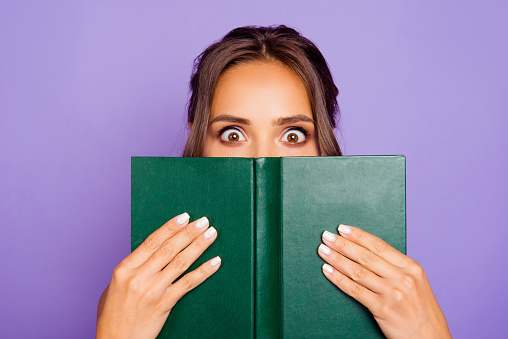 Close-up photo portrait of pretty sweet lovely smart clever cute wondered charming bookworm closing covering face with notebook notepad holding hands isolated violet background.