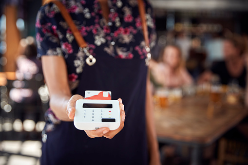 Close Up Of Waitress Holding Credit Card Payment Terminal In Busy Bar Restaurant