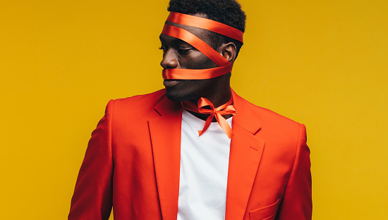 African man with satin ribbon wrapped on his face against yellow background. Man in orange blazer rapped as a present.