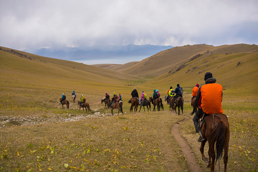 Back view of tourists on horseback riding tour, on the way to Song Kol Lake in Kyrgyzstan, which is in background. . The grassland is all around on cloudy and rainy day. It is moment of three days horseback riding, trekking over mountains from Kochkor to The Lake Song-Kul.