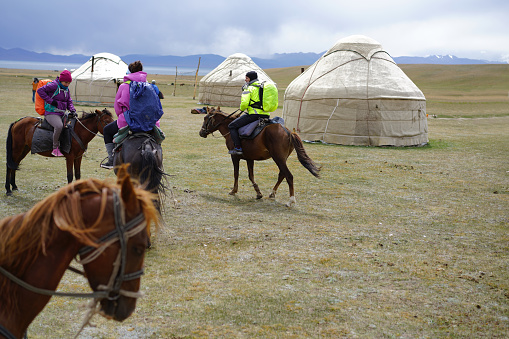 A tourists on horseback riding on rain, passing yurts and approaching Song Kol Lake in Kyrgyzstan on grassland and in background mountain ranges with snow and cloudy sky. It is moment of three days horseback riding, trekking over mountains from Kochkor to The Lake Song-Kul.