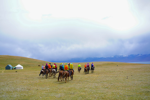 A big group of tourists on horseback riding passing by Yurts and approaching Song Kol Lake in Kyrgyzstan with grassland and in background mountain ranges with snow and cloudy sky. It is moment of three days horseback riding, trekking over mountains from Kochkor to The Lake Song-Kul.