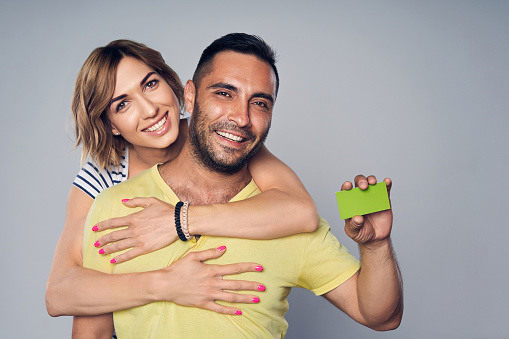Happy couple embracing, looking at camera showing blank credit card, over grey background