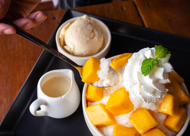 Woman use spoon take shaved ice dessert, served with mango sliced.  Served with vanilla ice cream and whipped cream.