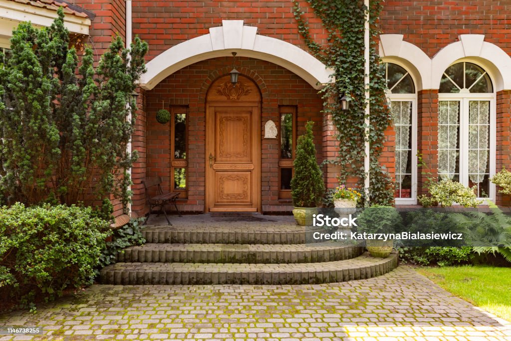 Plants, bushes and trees in front of wooden door of red brick mansion with windows Architecture Stock Photo