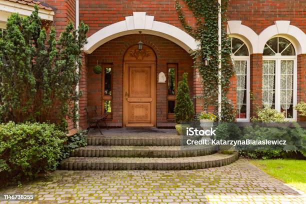 Plants Bushes And Trees In Front Of Wooden Door Of Red Brick Mansion With Windows Stock Photo - Download Image Now