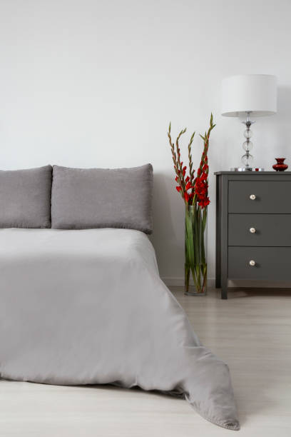 istock Red flowers in glass vase between king size bed with grey bedding and grey commode with elegant lamp on it 1146738295