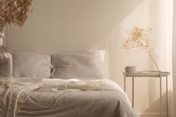 Small bedside table with glass vase and coffee cup next to big comfortable king size bed with white bedding, real photo