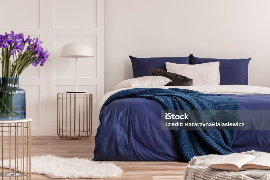 Purple flowers in blue glass vase on stylish table in white bedroom interior with comfortable bed Purple Stock Photo