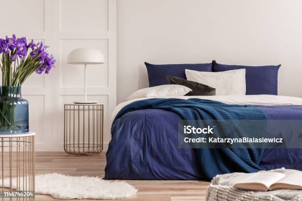 Purple Flowers In Blue Glass Vase On Stylish Table In White Bedroom Interior With Comfortable Bed Stock Photo - Download Image Now