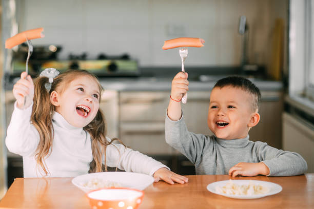 niño y niña niños en la cocina comiendo salchichas con pasta es muy divertido y amable - healthy eating snack child domestic kitchen fotografías e imágenes de stock